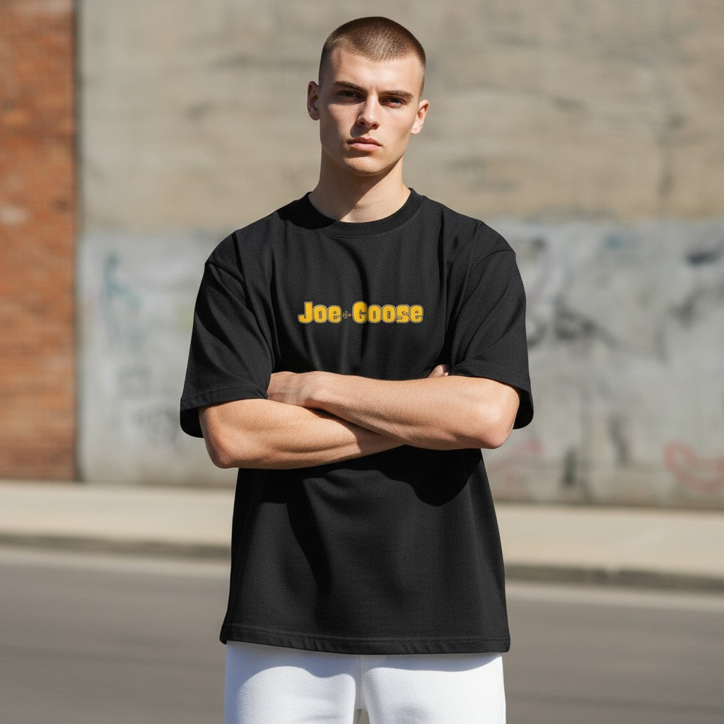 Model wearing a black Joe the Goose Apparel heavy weight cotton tee with yellow logo in an urban street setting.