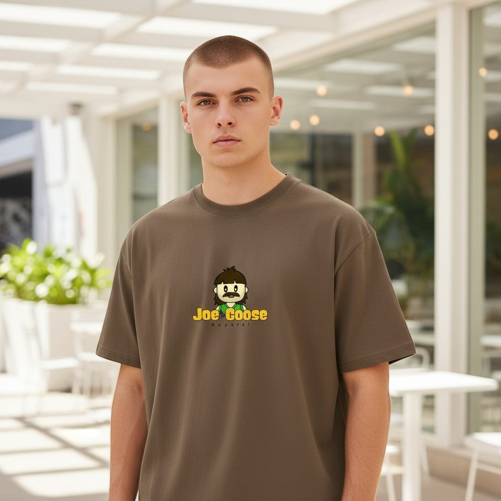 A man wearing a Joe the Goose Apparel brown heavy cotton t-shirt with a cartoon logo in a bright outdoor setting.
