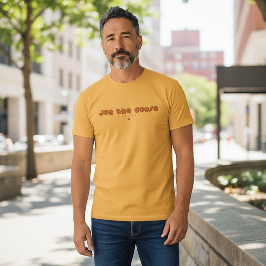 A man wearing a mustard yellow Joe the Goose Apparel classic cotton t-shirt with a retro logo, standing outdoors.