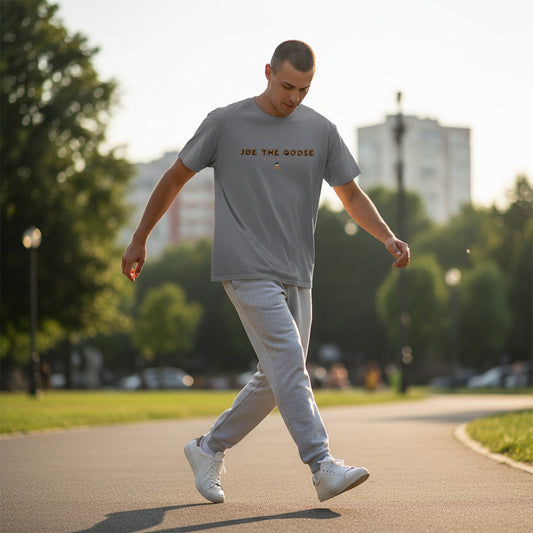 Man wearing a grey Joe the Goose Apparel cotton t-shirt with neon logo, walking in a park at sunset.