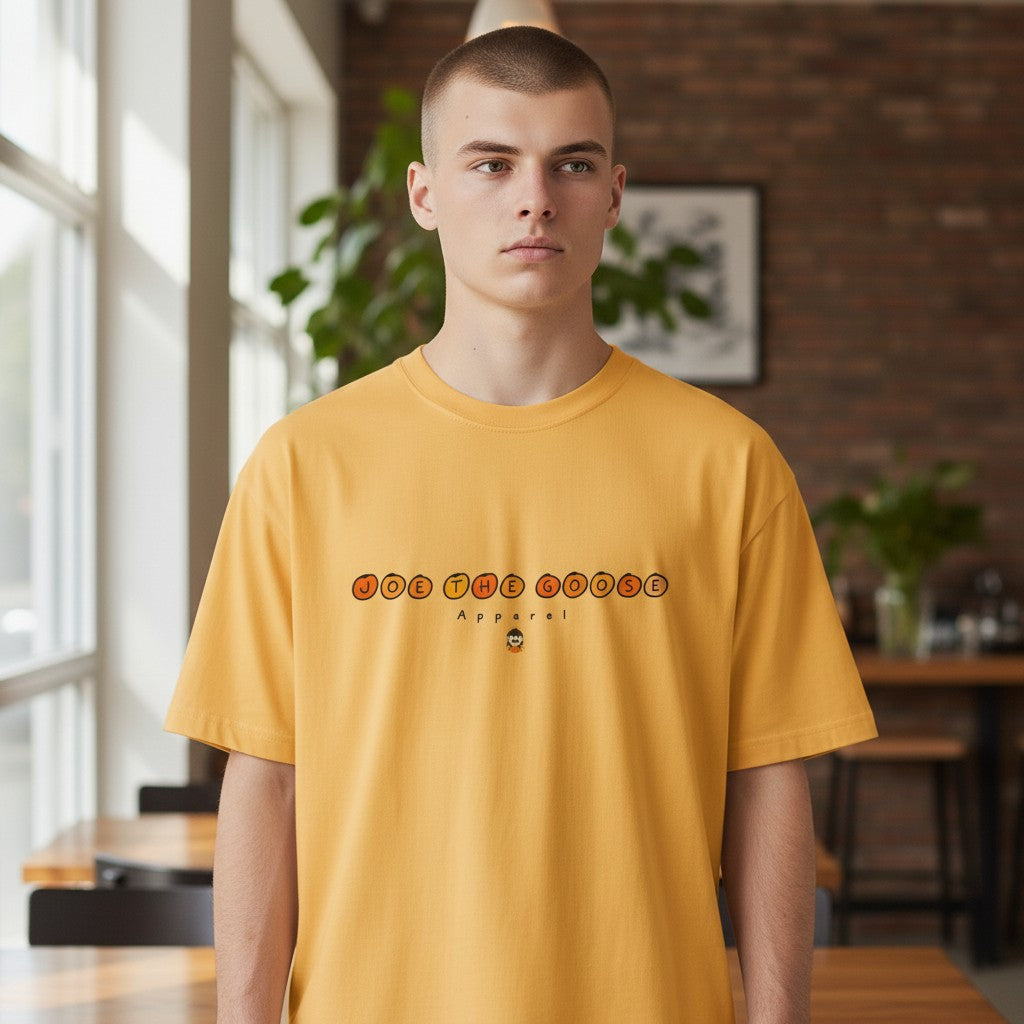Man wearing a yellow Joe the Goose Apparel classic cotton t-shirt with a circular logo graphic in a cafe setting.